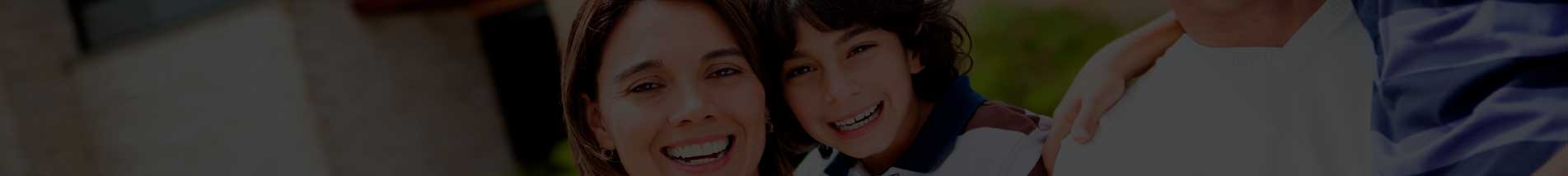Mujer y niño sonrientes, el niño con frenillos, abrazados junto a otra persona sobre un fondo exterior desenfocado.