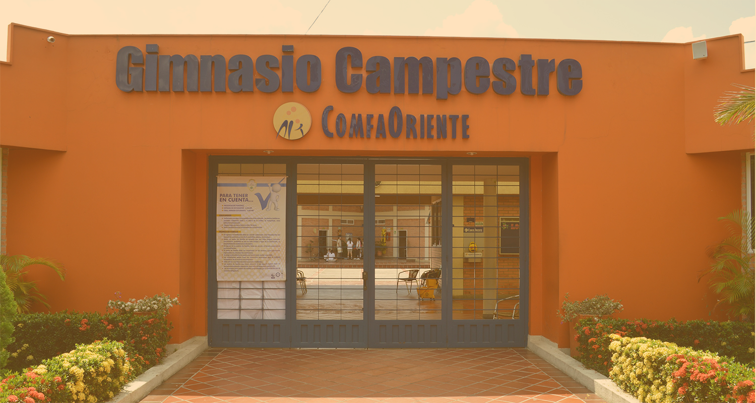Fachada naranja del Gimnasio Campestre ComfaOriente, letrero grande, puertas de vidrio con rejas y jardineras floridas.