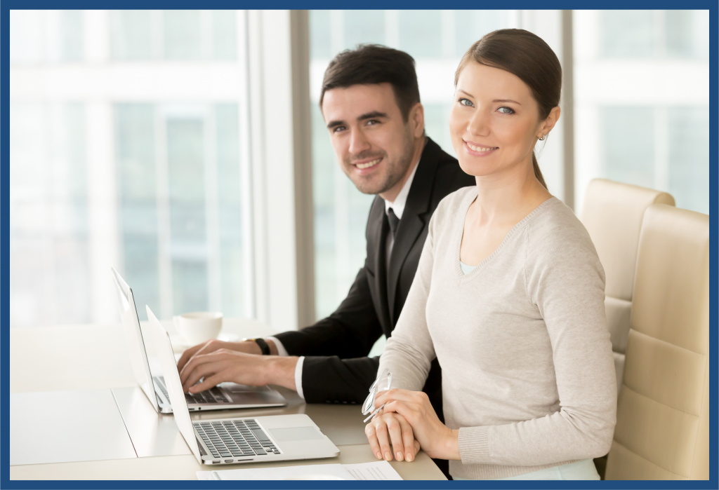 Dos profesionales sonrientes en oficina junto a ventanas; mujer con suéter claro sostiene gafas y hombre en traje usa laptop.
