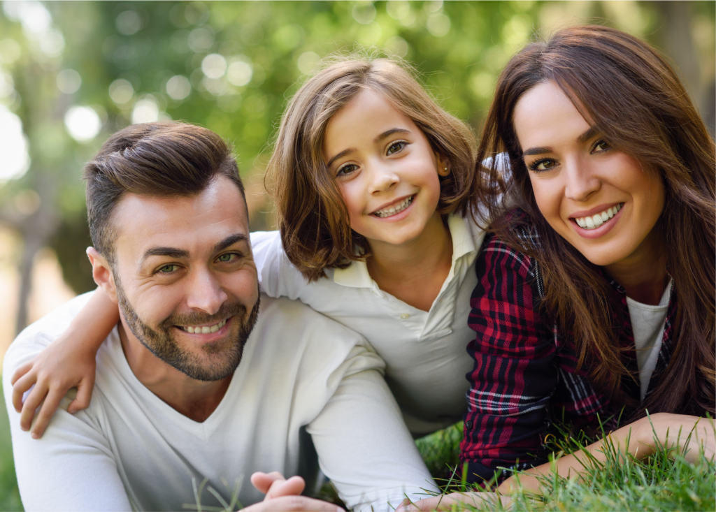 Padres y su hija sonriendo recostados sobre el césped, niña al centro con cabello castaño y padres mirando a cámara.