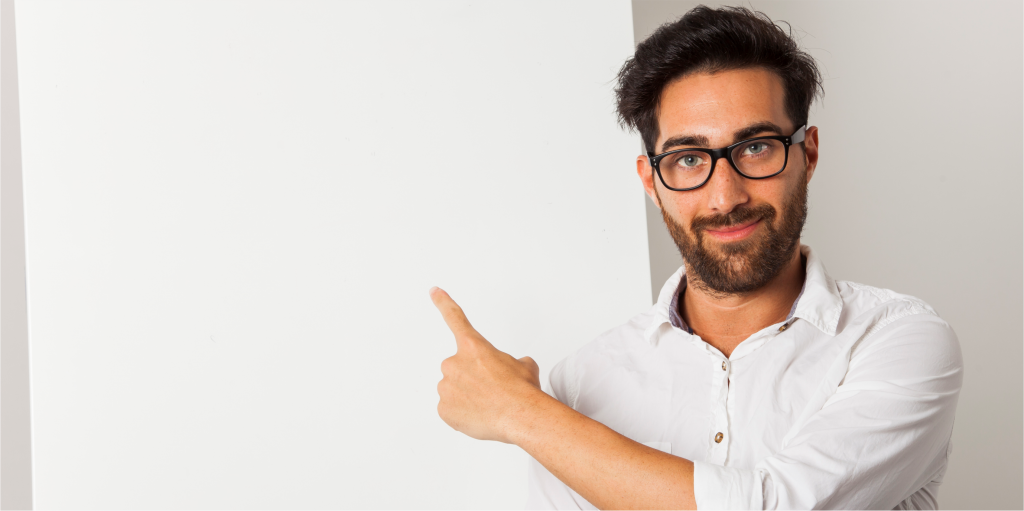 Hombre con gafas y barba, camisa blanca, sonriendo y apuntando con el dedo hacia un panel blanco vacío.