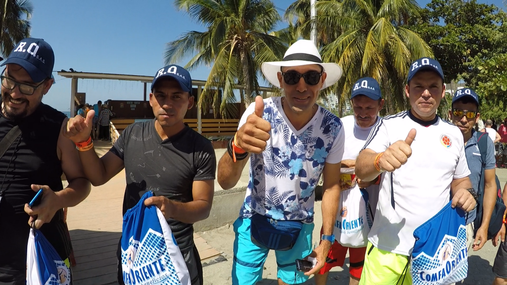 Seis hombres frente a palmeras en la playa, el del centro con sombrero blanco y camisa floral mostrando pulgar arriba.