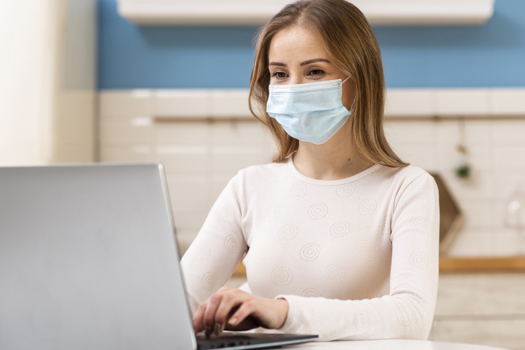 Mujer con mascarilla quirúrgica usando una laptop en una cocina, vestida con blusa blanca de patrones circulares.