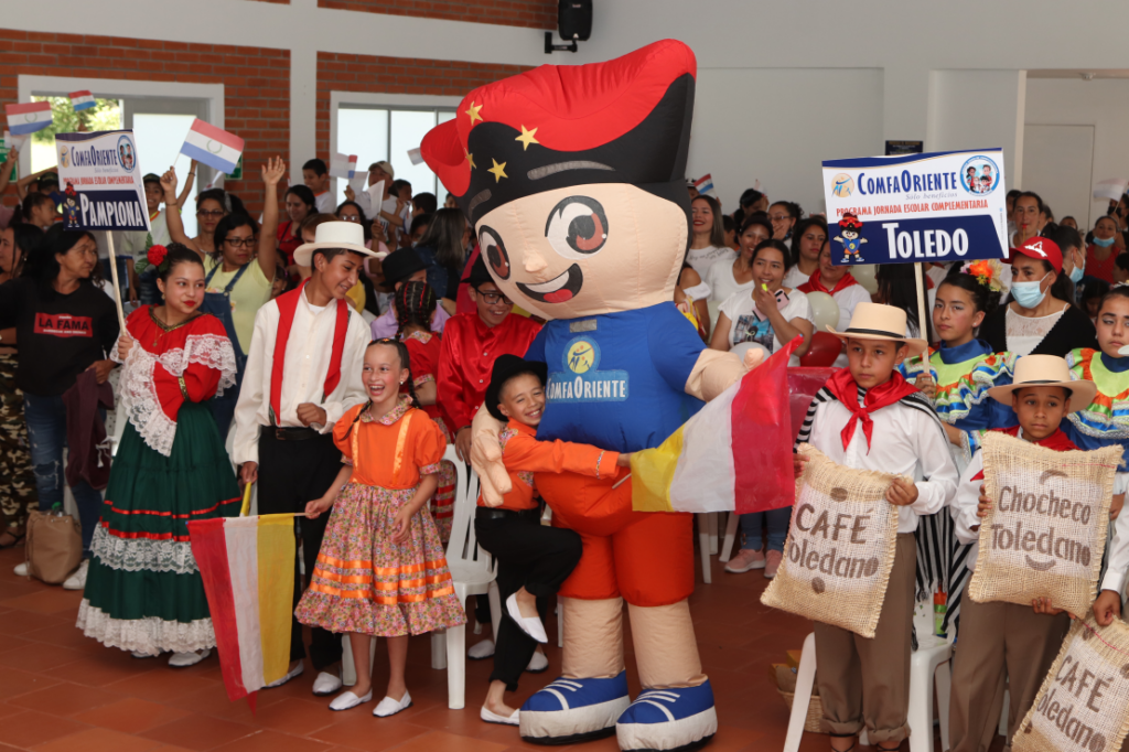 Niños en trajes tradicionales con una gran mascota de ComfaOriente, banderas y un cartel de Toledo en un acto interior.