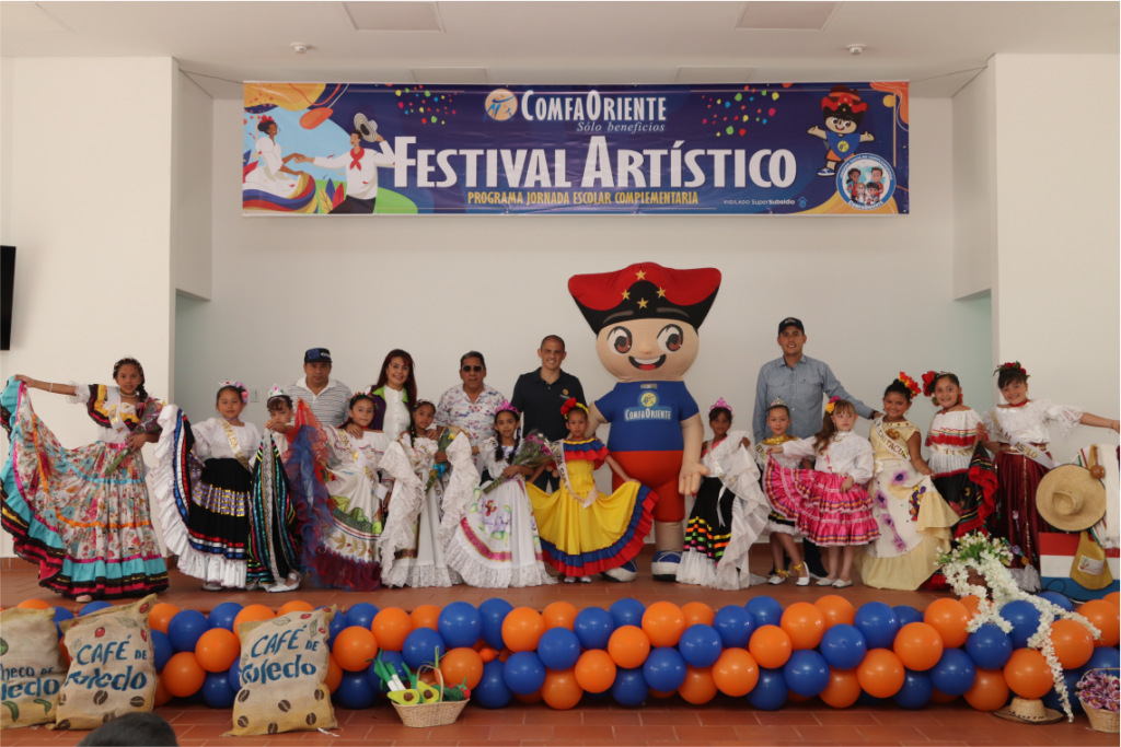 Niños y adultos en trajes folclóricos sobre un escenario bajo una pancarta Festival Artístico, con mascota inflable, globos y sacos de café