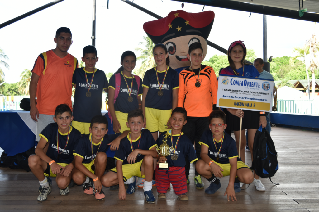 Equipo infantil con uniformes azul y amarillo, medallas y trofeo, posando junto a cartel ComfaOriente.