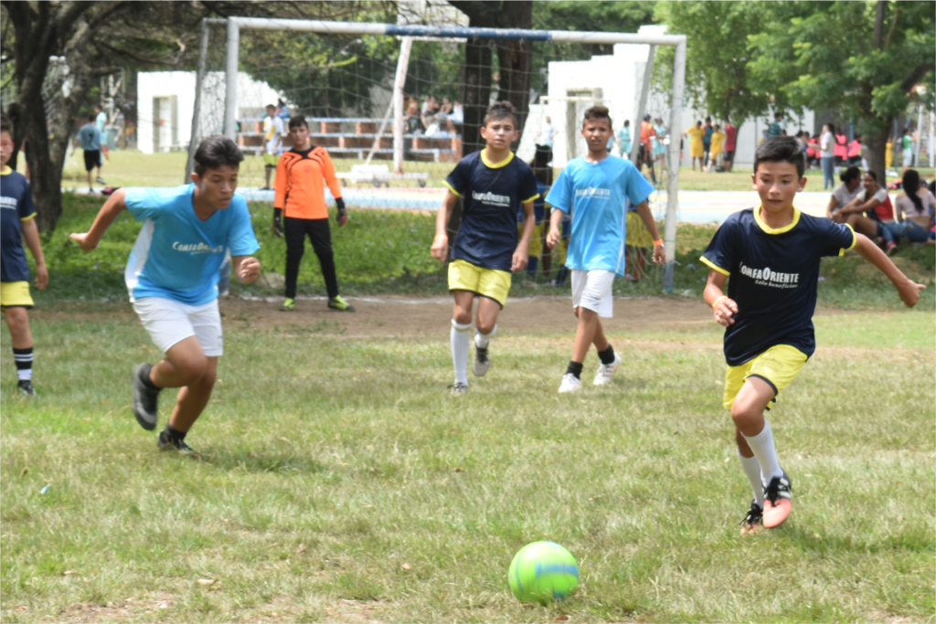 Niños en partido de fútbol: jugador con camiseta azul y short amarillo corre hacia un balón verde; portero en naranja