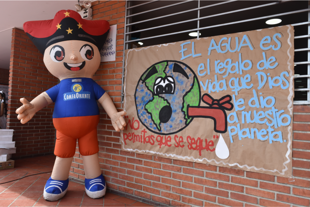 Muñeco inflable con camiseta Comfaoriente junto a cartel en ladrillo con dibujo del planeta y mensaje para cuidar el agua.