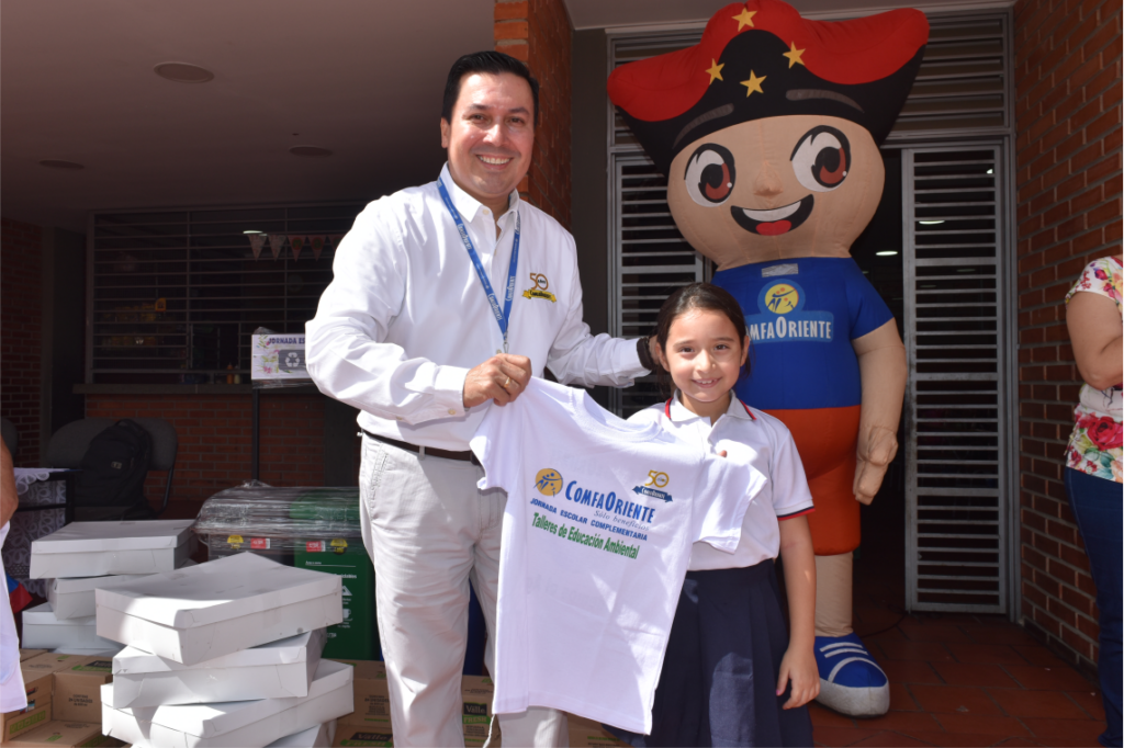 Hombre y niña sonrientes sostienen una camiseta blanca con el logo ComfaOriente y un personaje inflable detrás.