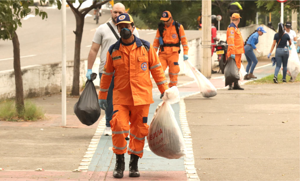 Persona con uniforme naranja y mascarilla cargando bolsas de basura en una vía peatonal, con más voluntarios al fondo.