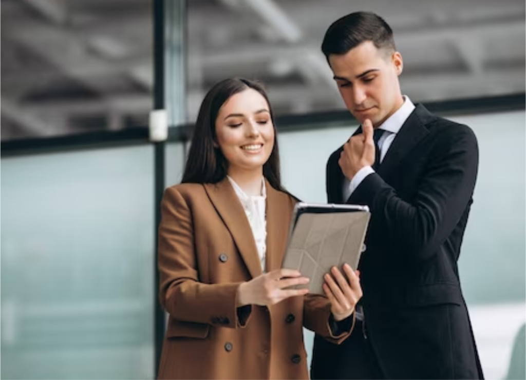 Mujer con blazer marrón y hombre de traje negro revisan una tableta juntos en oficina, ella sonríe y él parece pensativo.