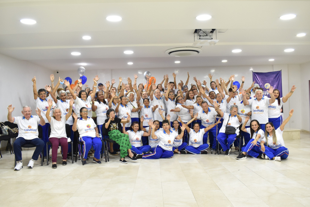Grupo grande de adultos mayores con uniformes blancos y pantalones azules, sonriendo y levantando las manos en salón iluminado.