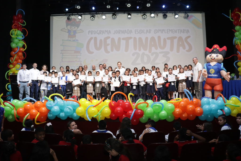 Niños en un escenario sosteniendo diplomas en la ceremonia Cuentinautas 2023, con decoración de globos y una mascota.