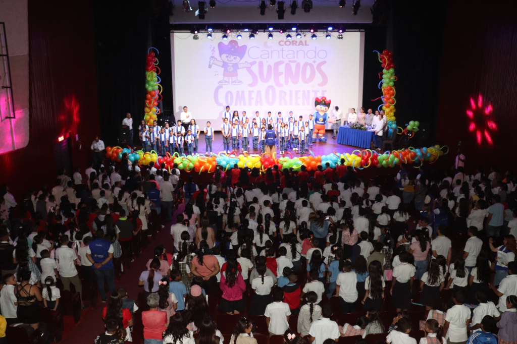 Niños en coro en un escenario decorado con globos multicolores y público numeroso en el auditorio