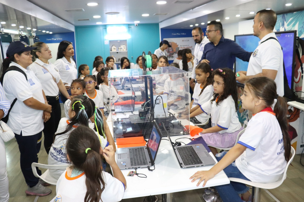 Grupo de niñas y niños con camisetas blancas observan una impresora 3D y laptops mientras instructores explican en un aula móvil.